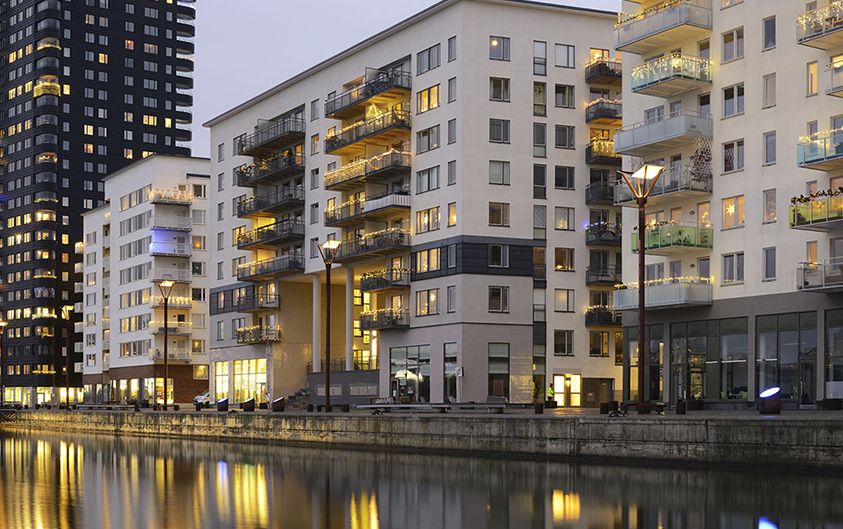 Moderne Wohngebäude am Wasser, die sich in einem ruhigen Kanal spiegeln, beleuchtet von Abendlichtern, mit der Skyline der Stadt im Hintergrund.