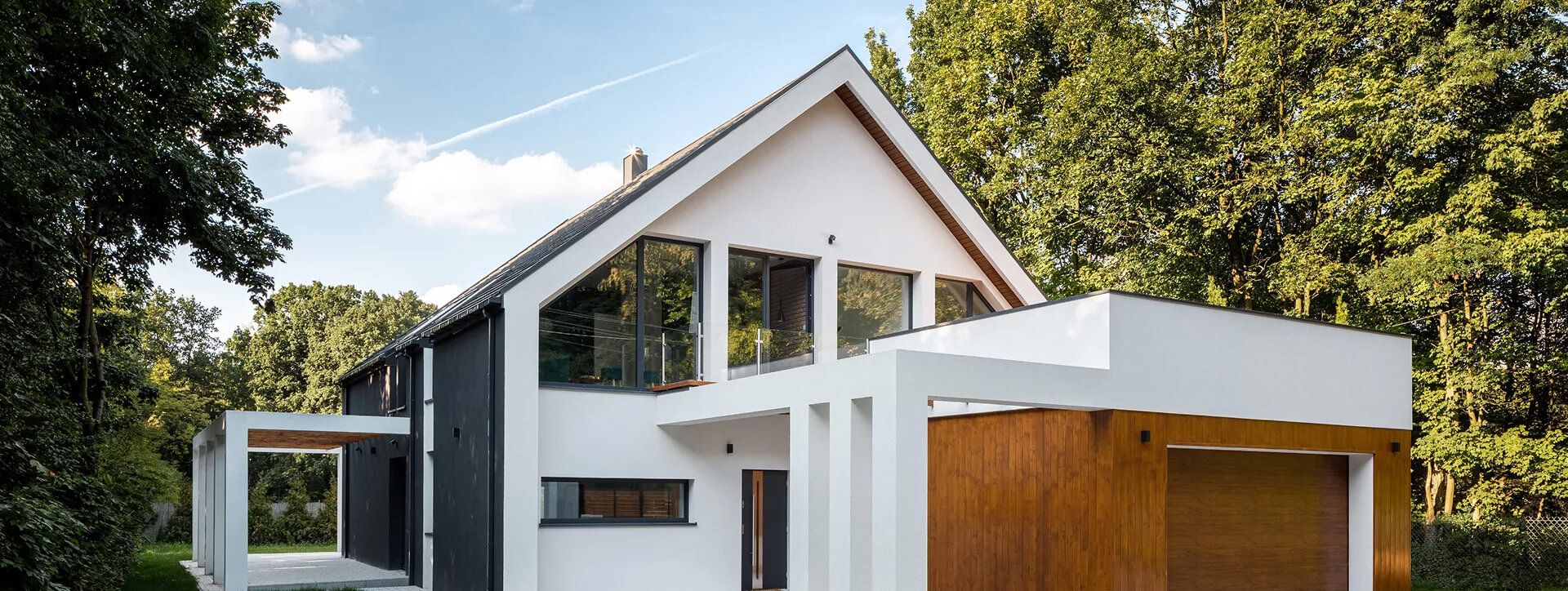Modernes zweistöckiges Haus mit einer Mischung aus weißer und dunkler Verkleidung, großen Fenstern und umgeben von üppig grünen Bäumen unter einem hellen Himmel.