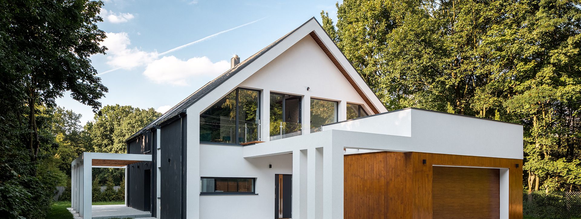Modernes zweistöckiges Haus mit einer Mischung aus weißer und dunkler Verkleidung, großen Fenstern und umgeben von üppig grünen Bäumen unter einem hellen Himmel.